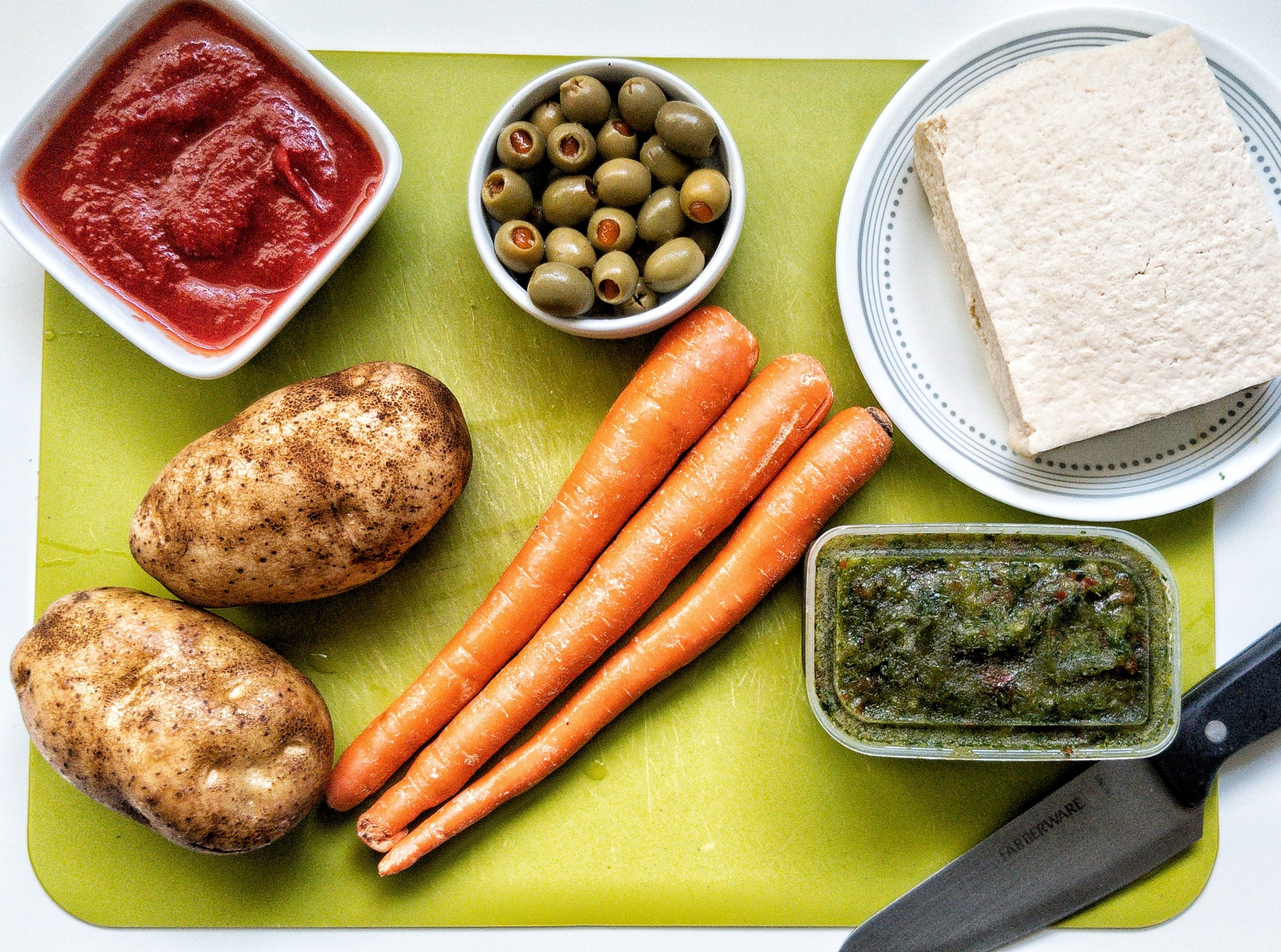 Green chopping board with whole potatoes, carrots, sofrito, tofu block, green Spanish olives, and tomato sauce