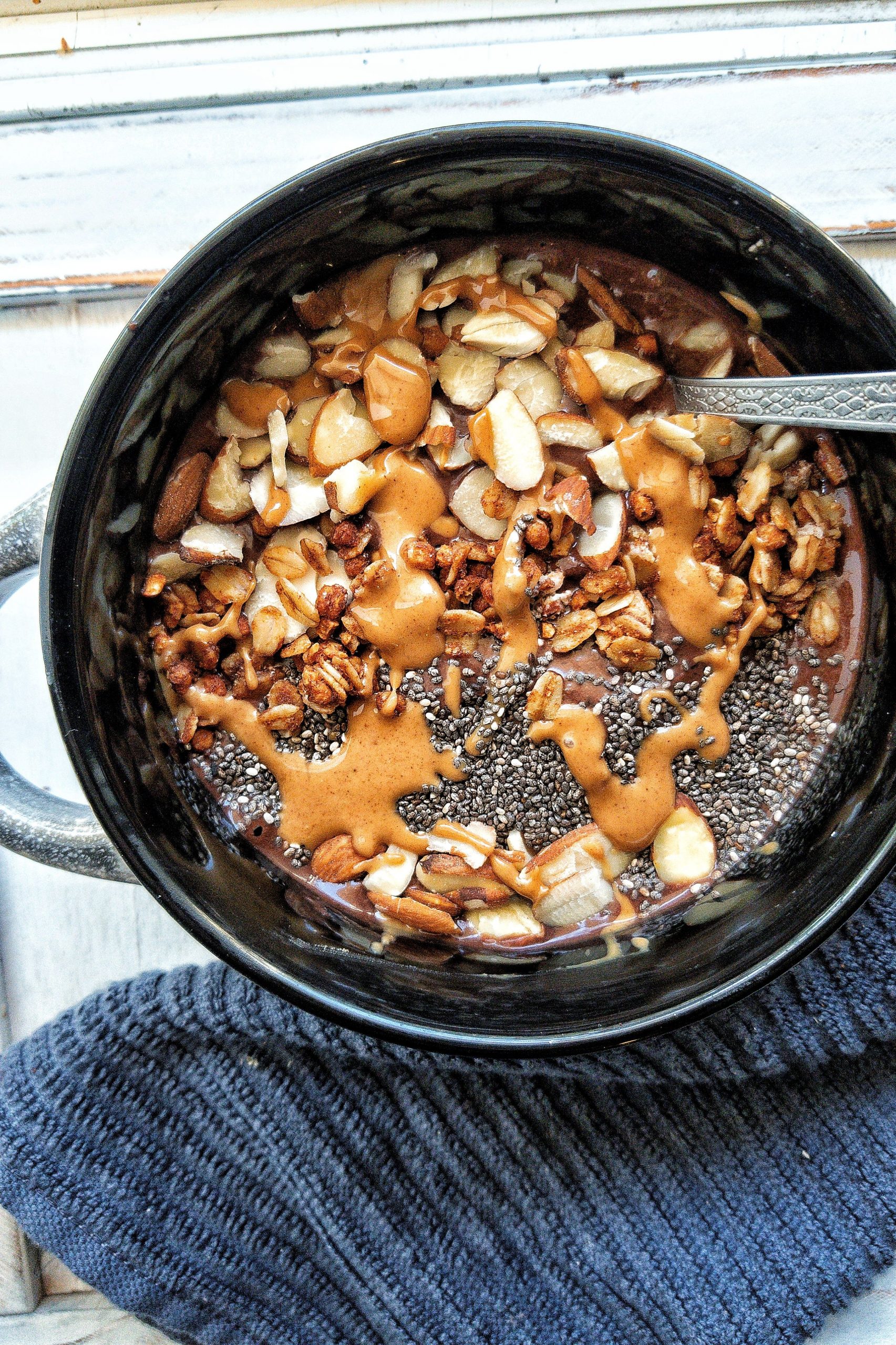 A chocolate banana smoothie bowl topped with peanut butter, chia seeds, sliced almonds, and granola