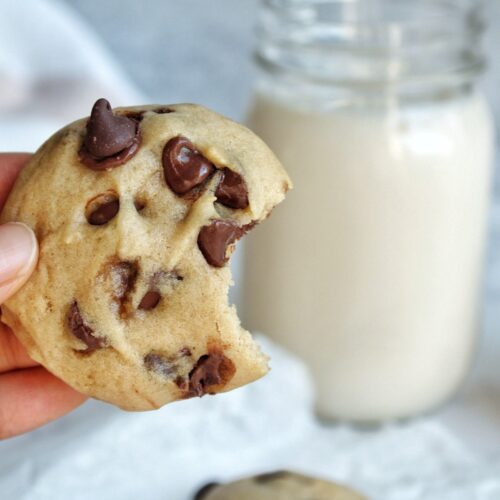Bitten chocolate chip cokies with a jar of milk and a stack of more cookies in the background.