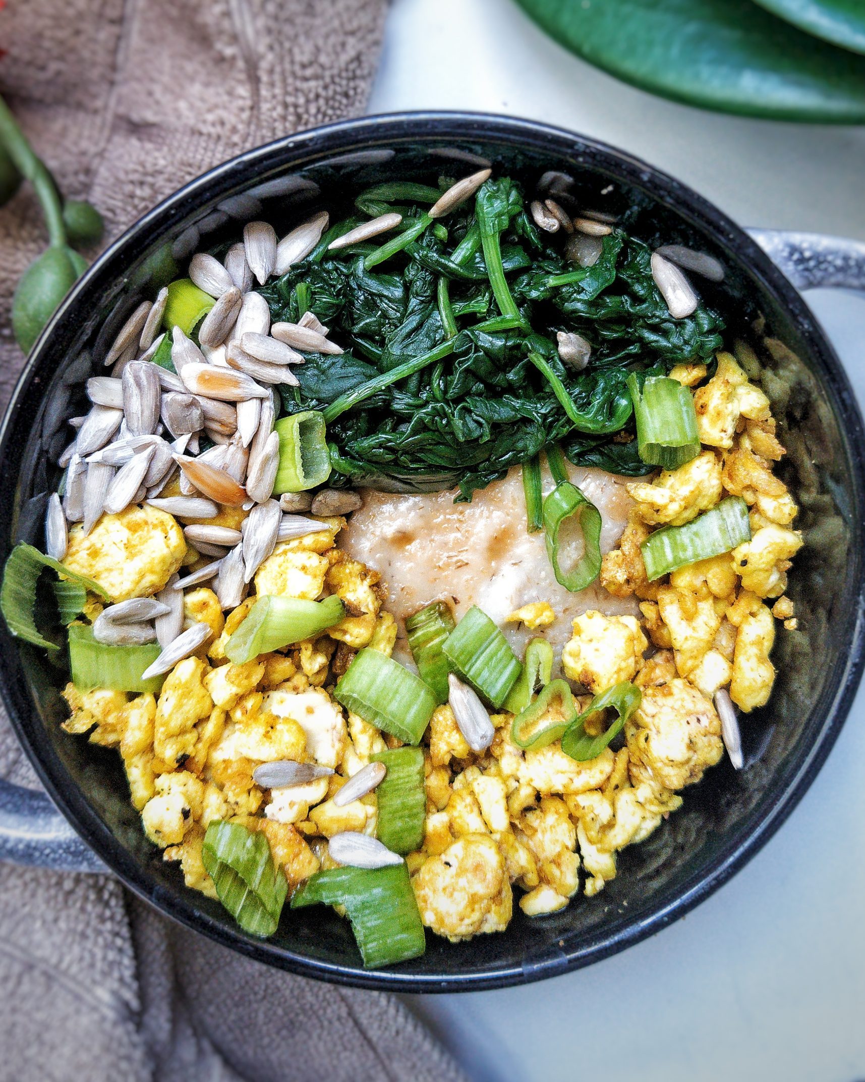 Bowl of creamy oat bran topped with tofu scramble, steamed spinach, sunflower seeds, chopped green onions, and a drizzle of soy sauce