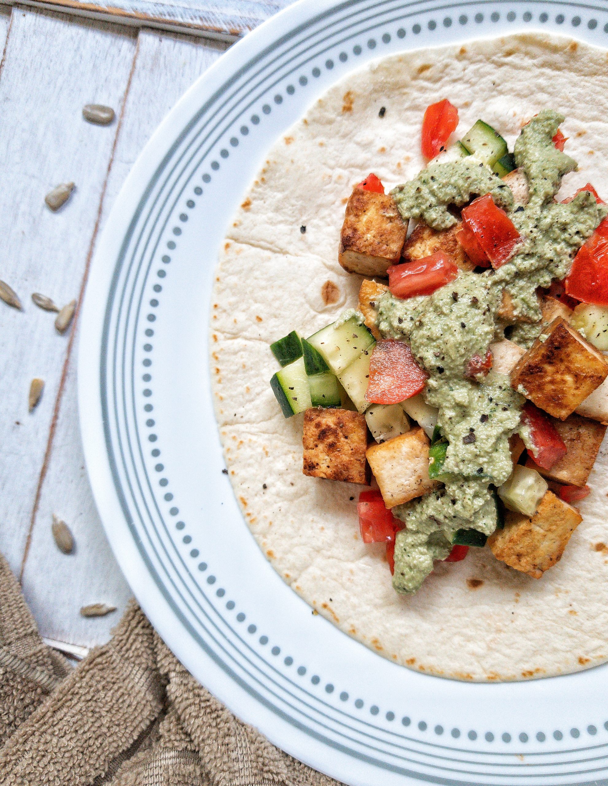 Flour tortilla laid flat and topped with tomatoes, pan-fried tofu, chopped cucumber, a drizzle of creamy cilantro sauce, and ground black pepper