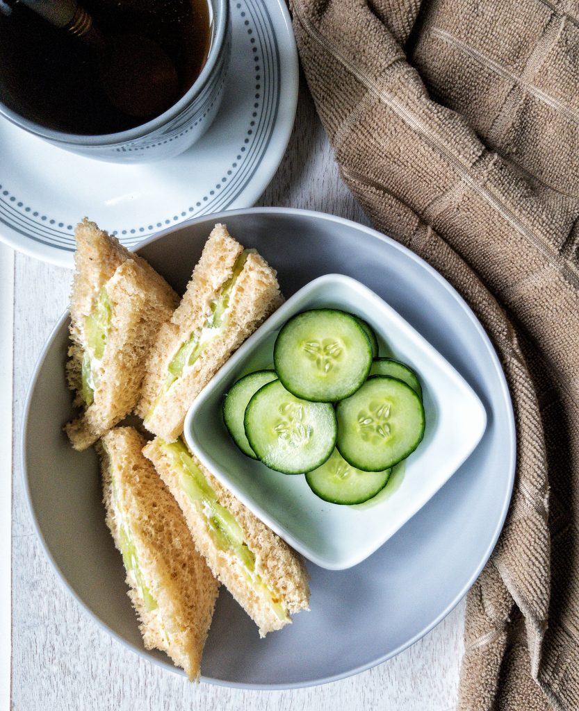 Four small cucumber tea sandwiches with a small side of sliced cucumbers alongside Earl Grey tea.