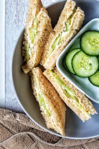 Closeup of cucumber tea sandwiches with a side of sliced cucumbers in a small, white dish