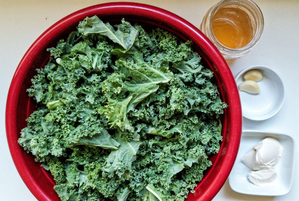 A bunch of raw kale in a large mixing bowl with measured vegan butter, garlic, and vegetable stock