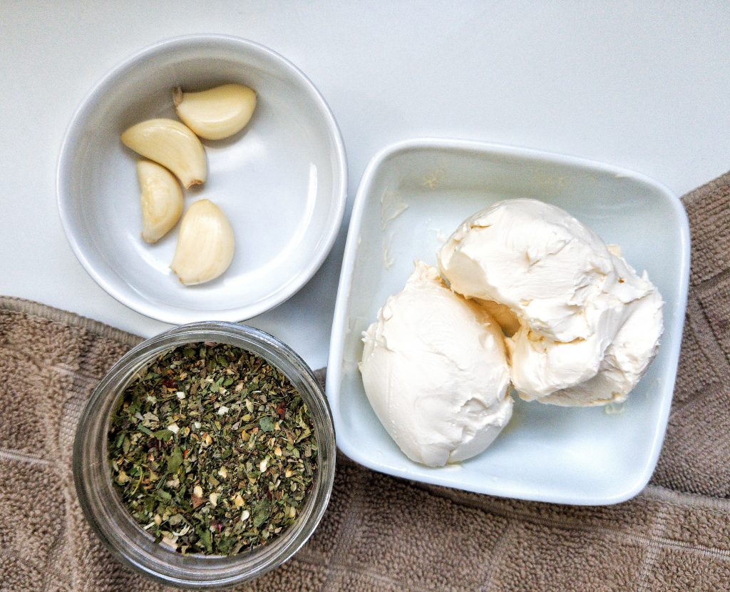 Whole garlic cloves, vegan butter, and Italian seasoning measured out in small bowls.