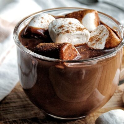 Creamy coconut milk hot chocolate with coffee in a clear glass mug. Topped with marshmallows, sprinkled with cinnamon.