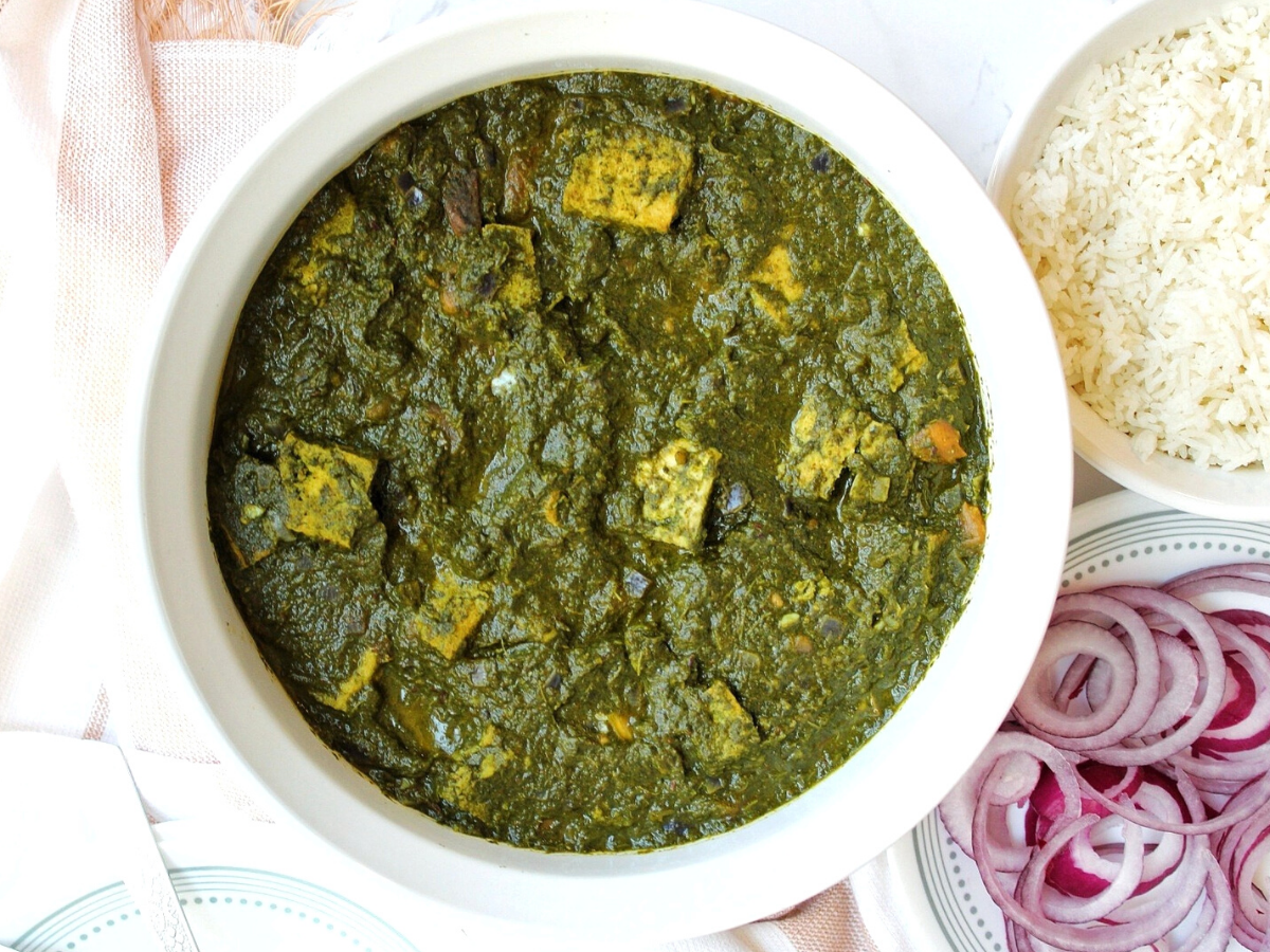 Vegan palak paneer in a large white bowl with a side of sliced onions and white rice.