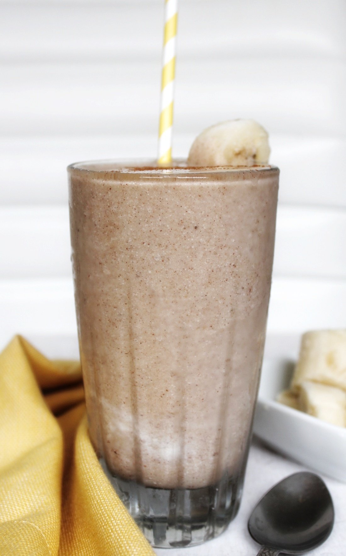 Milkshake in a glass cup with sliced banana and a yellow white stripped straw