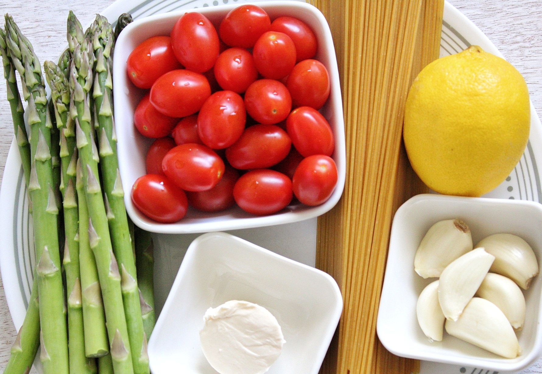 Uncooked spaghetti, whole lemon, garlic cloves, vegan butter, asparagus, and tomatoes on a plate 