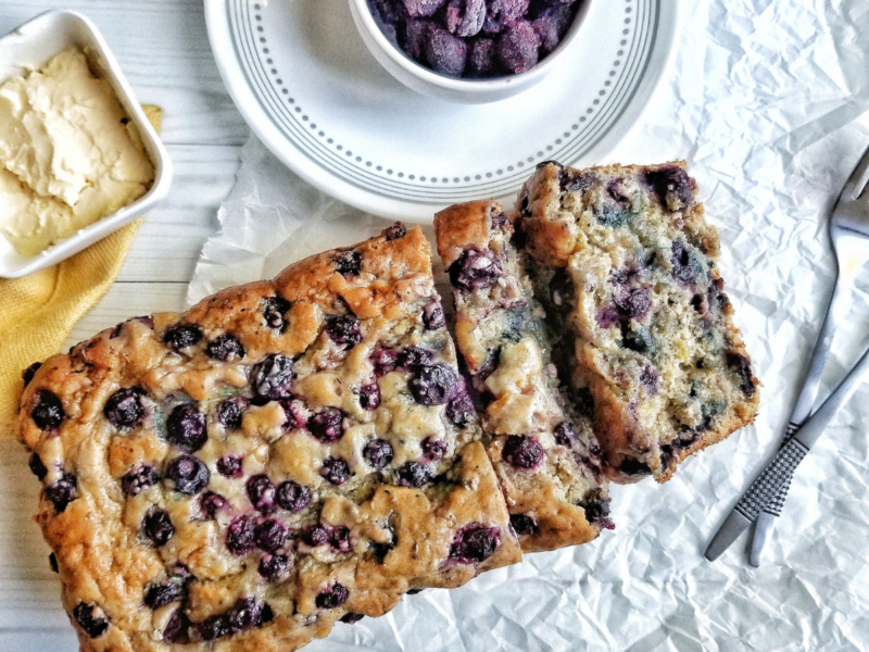 Blueberry banana bread with a bowl of frozen berries and vegan butter