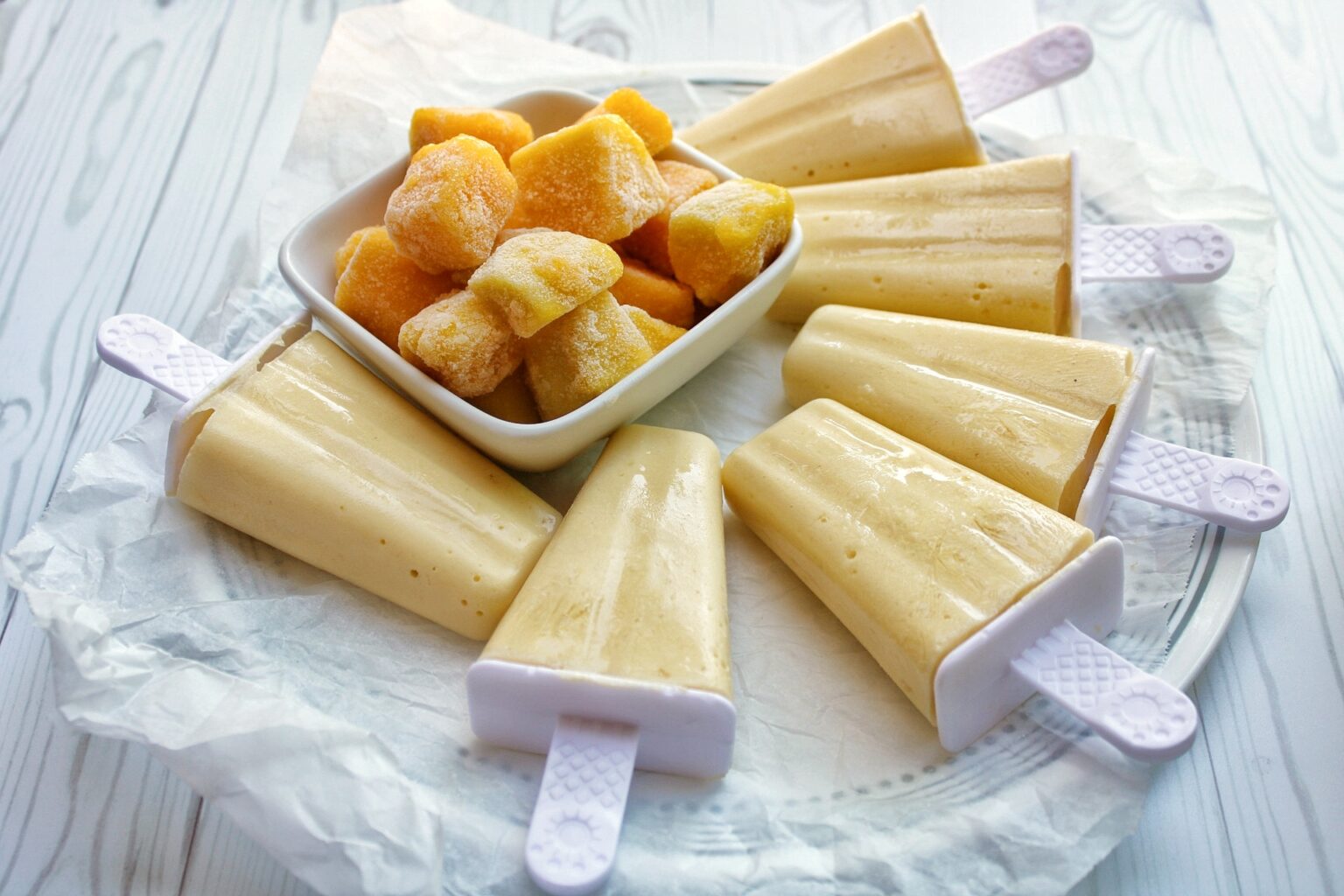 Six mango coconut ice pops on a plate with frozen mango in a bowl. 