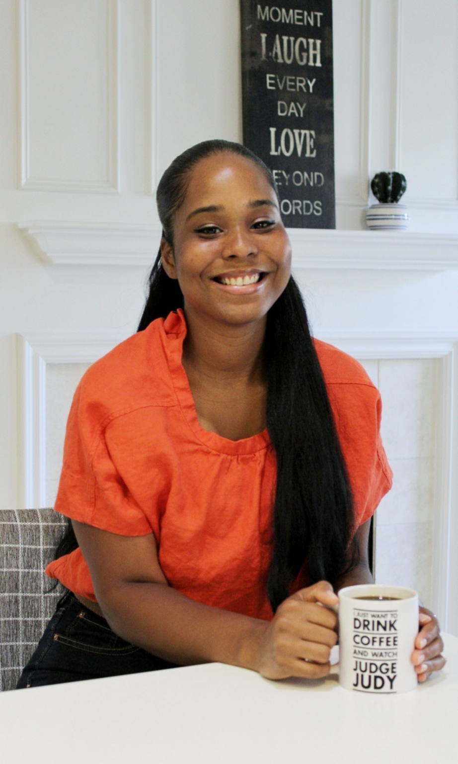 Aly in an orange blouse sitting at a kitchen island with a coffee cup that says "I just want to drink coffee and watch Judge Judy." 