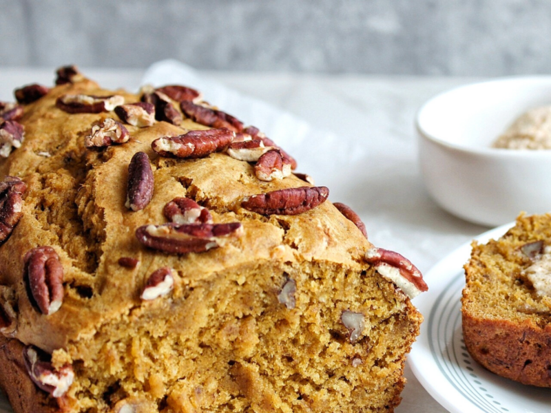 Loaf of pumpkin banana bread topped with choppedpecans with a slice on the side.