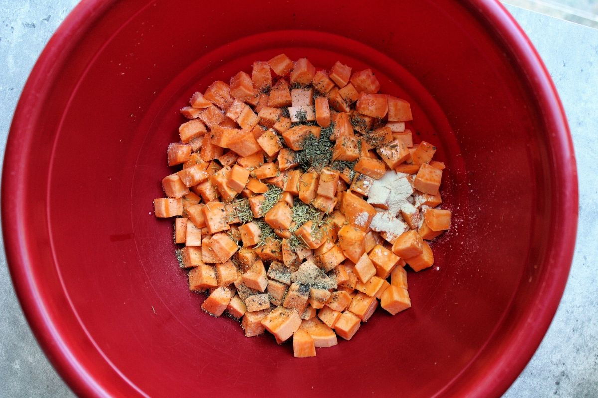 Bowl of uncooked sweet potato cubes with unmixed olive oil, herbs, and spices in a red bowl. 