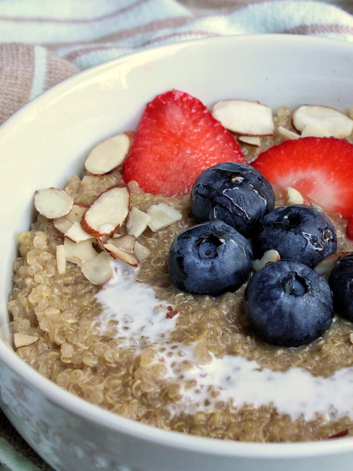 Bowl of quinoa porridge topped with blueberries, strawberries, sliced almonds, and a drizzle of coconut milk. 