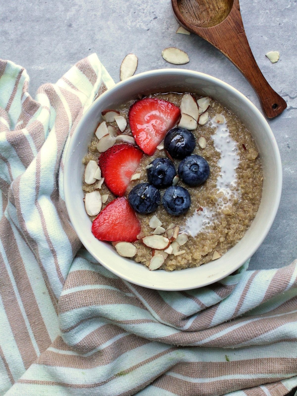 Qunioa porridge topped with strawberries, blueberries, sliced almonds, and coconut milk. With a wooden spoon and a kitchen towel. 