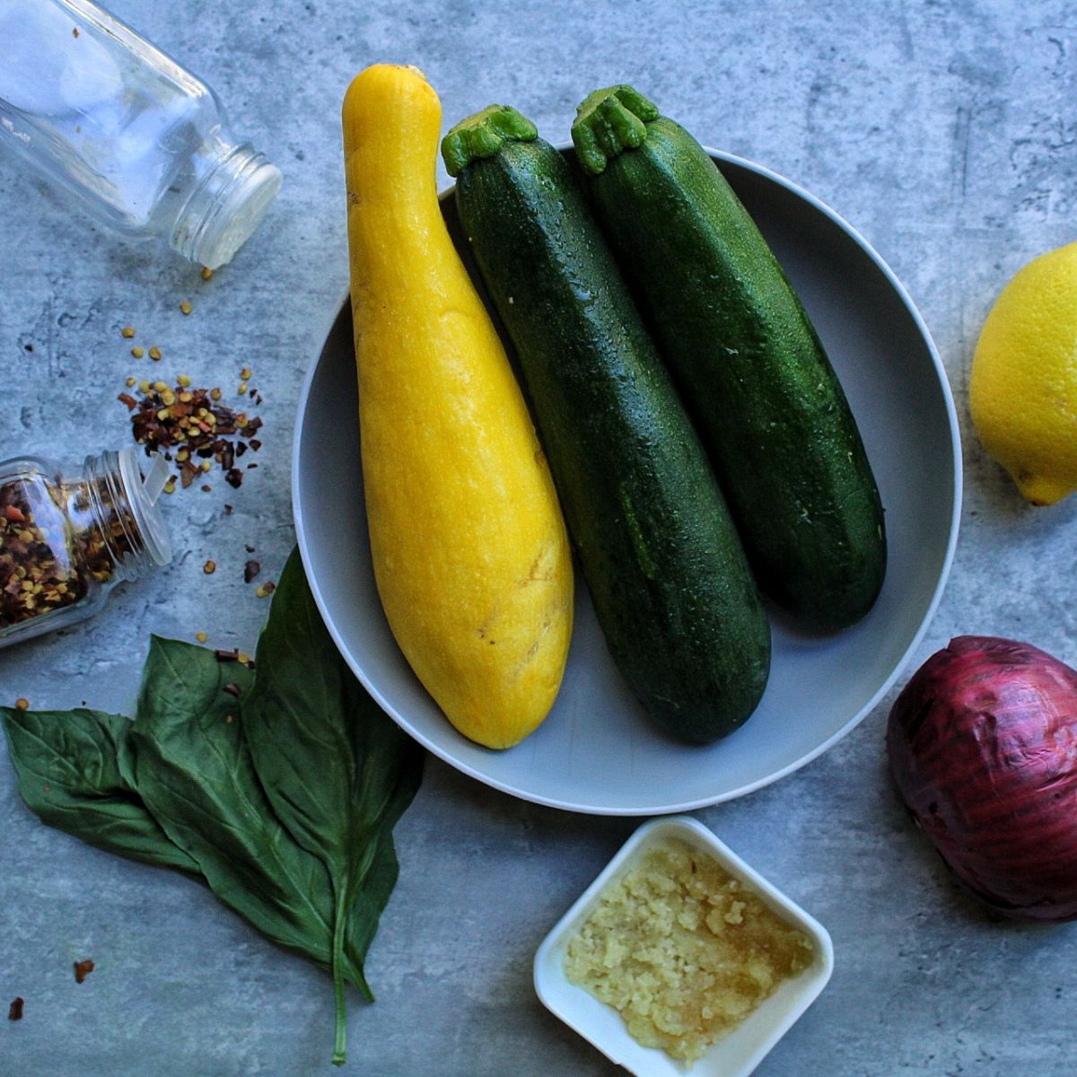 Yellow and green zucchini in a gray bowl, salt shaker, red pepper flakes shaker, whole basil leaves, minced garlic, red onion, and a lemon. 