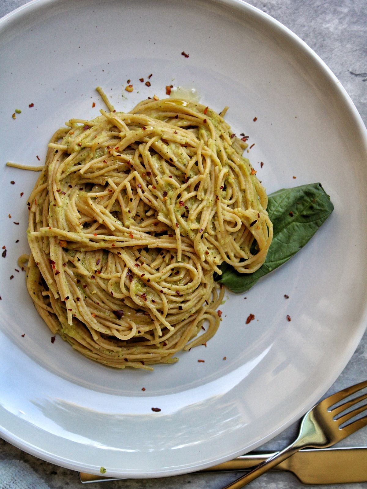 Whole wheat spaghetti with zucchini pasta sauce, garnished with basil leaf and cracked red pepper all on a white plate with silverware. 