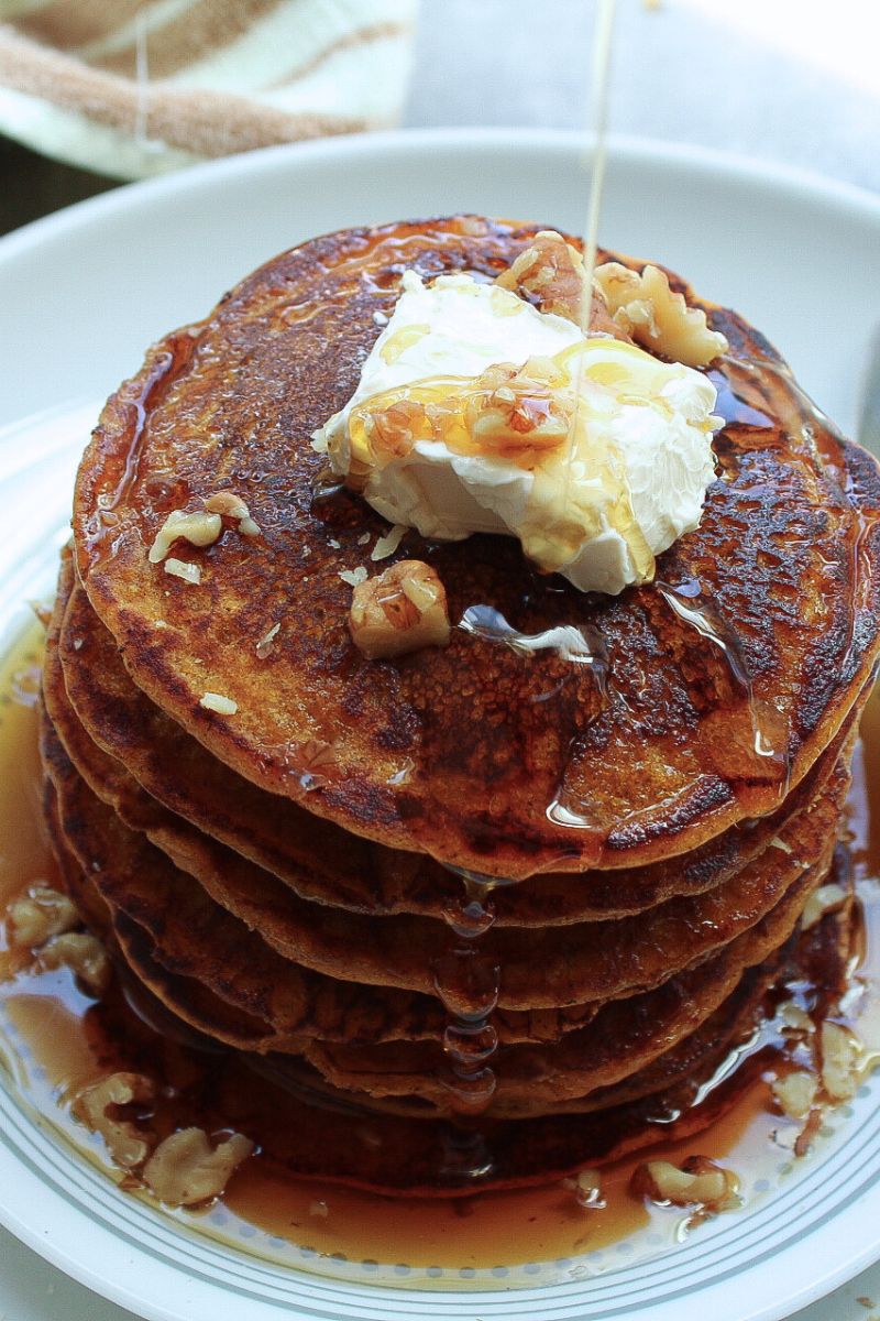Stack of vegan pumpkin pancakes with a dollop of vegan butter and a pour of maple syrup. Topped with chopped walnuts. 
