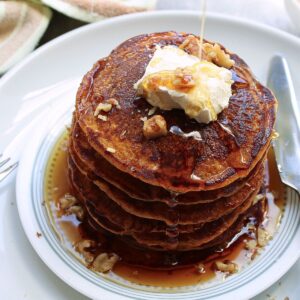 Vegan pumpkin pancakes with a pour of maple syrup, topped with vegan butter and chopped walnuts.