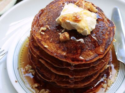 Vegan pumpkin pancakes with a pour of maple syrup, topped with vegan butter and chopped walnuts.